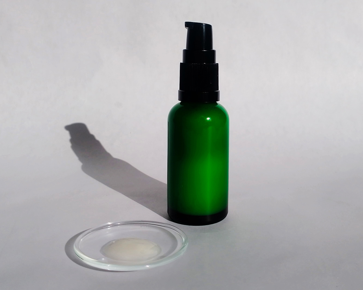 Sérum na tvár a fúzy v zelenej sklenenej fľaštičke a trochou séra na prihľadnom sklenenom podnose na bielom pozadí.